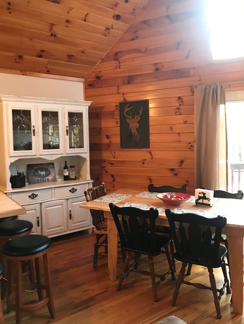 Rustic log cabin interior with wooden walls, white cabinet, dining table with black chairs, and deer artwork on wall