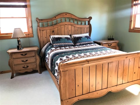 Bedroom with wooden bed frame, blue patterned bedding, nightstand with lamp, and light blue walls with windows