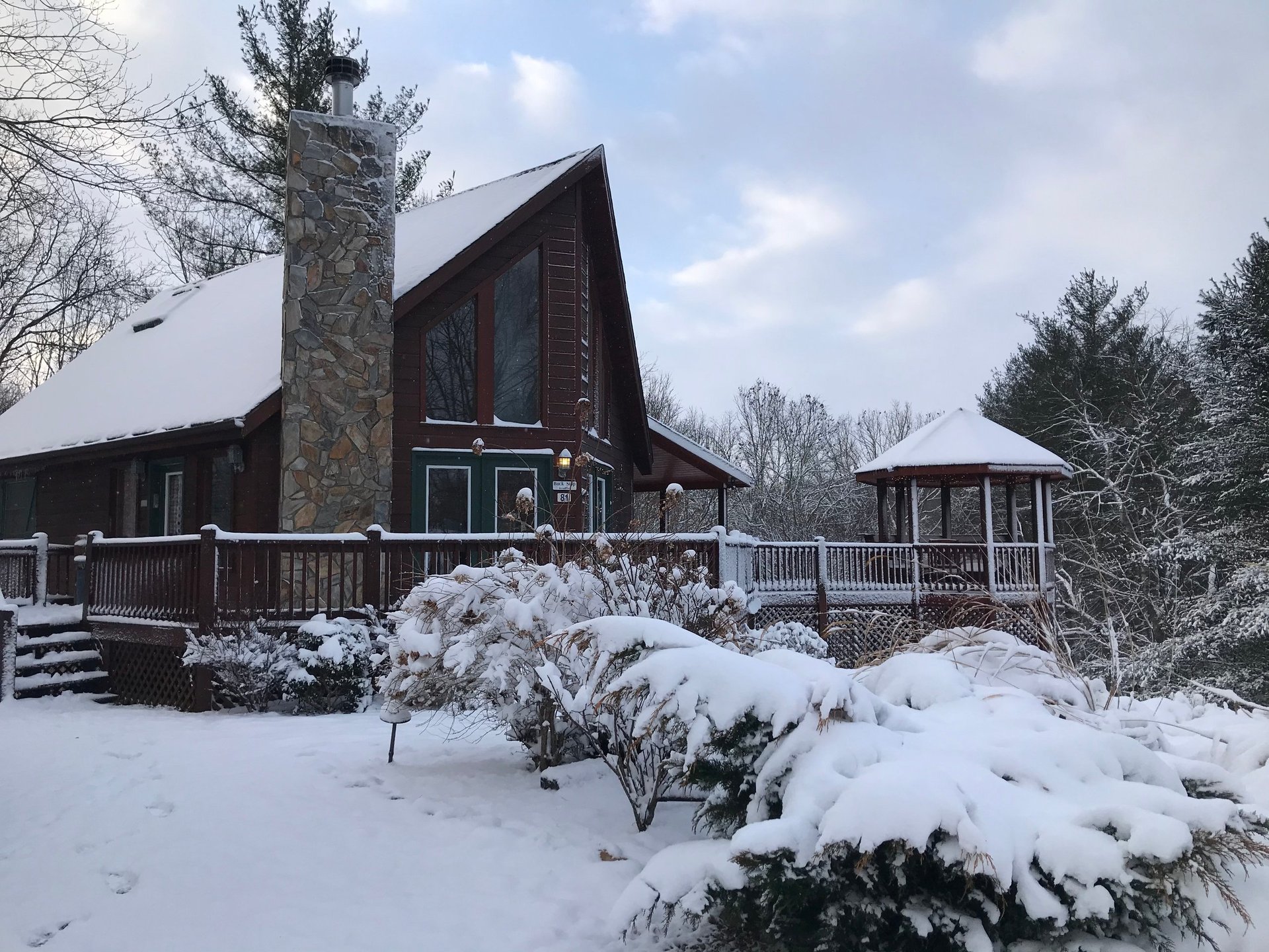 A-frame cabin with stone chimney surrounded by snow-covered evergreens and bare trees in winter