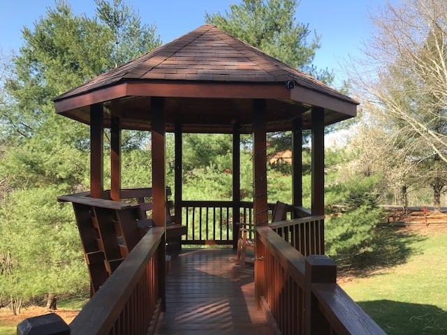 Wooden gazebo with peaked roof and railing on a deck, surrounded by green trees and grass