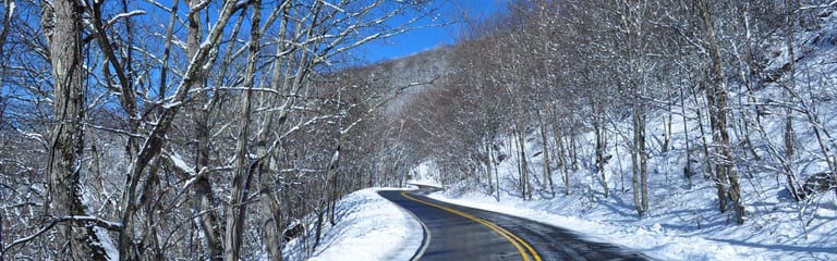 Snow-covered mountain road winding through winter forest with bare trees under blue sky