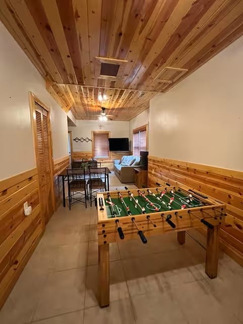Wooden rec room with foosball table in foreground, bar seating, TV, and wood-paneled ceiling