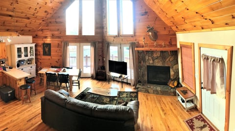 Interior of a rustic wooden cabin with open living space, black leather sofa, fireplace, and tall windows.