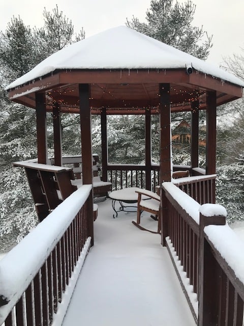 Snow-covered wooden gazebo with dark brown railings and seating, surrounded by snow-laden trees and bare branches