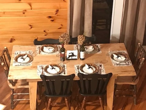 Wooden dining table set with five place settings, black chairs, decorative centerpieces, and beige curtains in a rustic log cabin interior