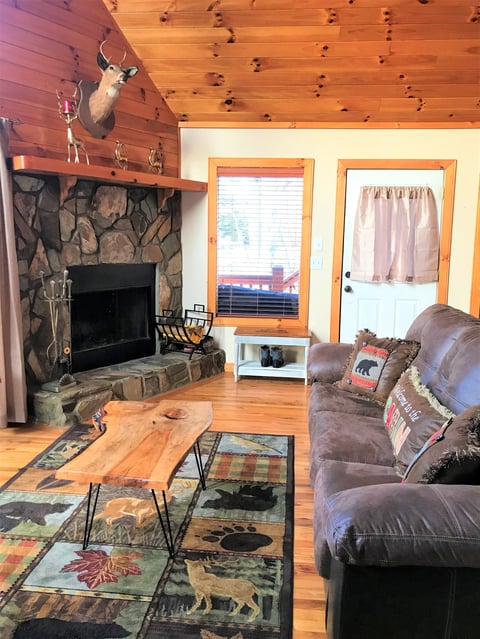 Rustic cabin living room with stone fireplace, mounted deer head, wood ceiling, leather seating, and natural wood coffee table